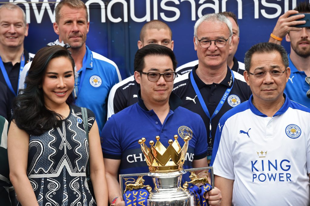 Leicester City's owner Vichai Srivaddhanaprabha (R) looks on as his son and vice-chairman Aiyawatt "Top" Srivaddhanaprabha (C) holds the Premier League trophy while posing for pictures ahead of a parade in an open-bus with the club's football players through Bangkok on May 19, 2016. Newly crowned English Premier League champions Leicester City received a royal seal of approval on May 19 at Bangkok's Grand Palace, with the Thai-owned team presenting its trophy to a portrait of the king before a bus parade through the capital. / AFP PHOTO / CHRISTOPHE ARCHAMBAULT