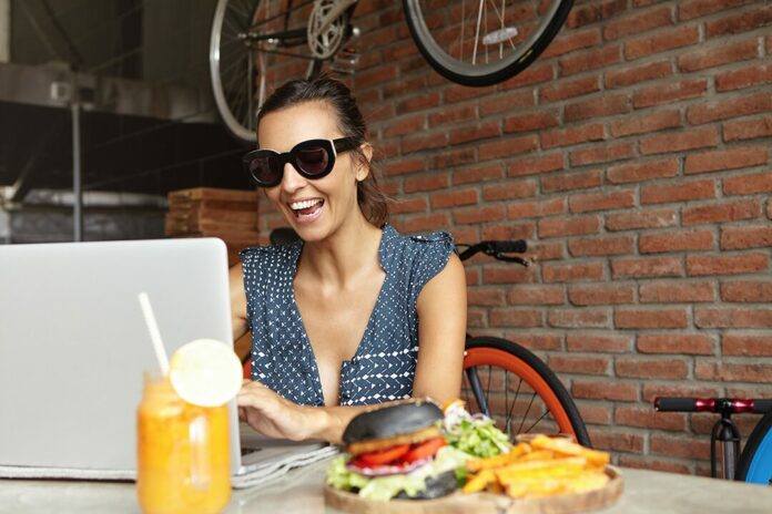 Cheerful woman blogger in trendy sunglasses recording webcam video of herself to post it on her blog, using wireless internet connection, sitting at cafe table with fresh orange juice and food